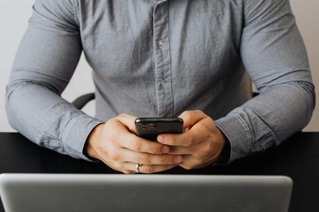 A person wearing a gray shirt holds a smartphone in front of a laptop, possibly at work.